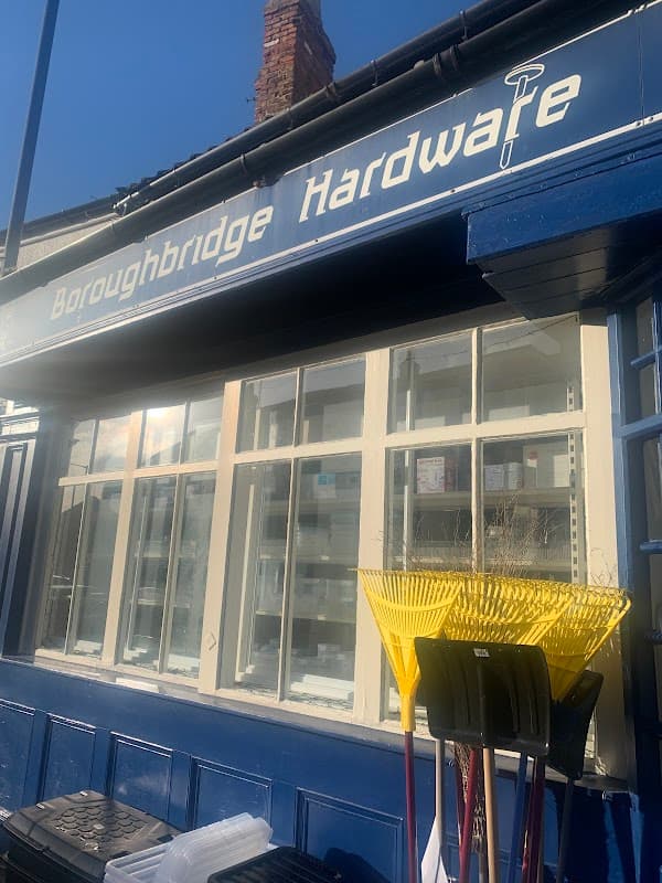 Sign for Boroughbridge Hardware Ltd with blue facade and yellow garden rakes displayed outside the window.