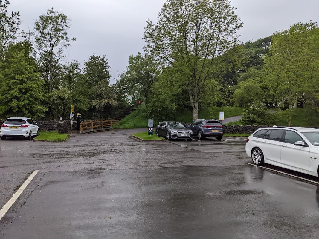 Pod Point Charging Station in a wet, tree-lined parking area with several parked cars and a footpath in the background.
