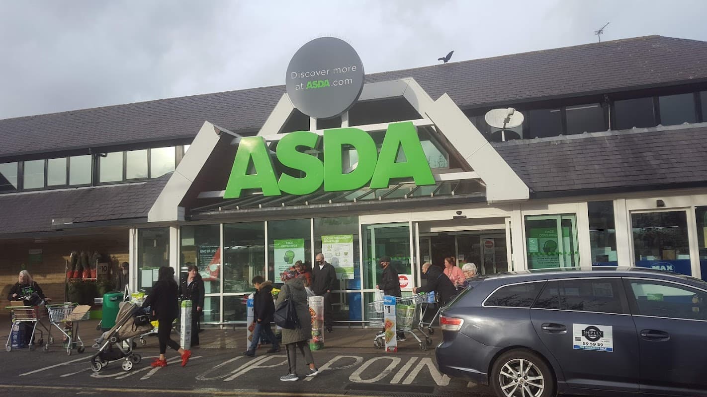 ASDA Superstore entrance with shoppers, carts, and a gray car parked nearby under a cloudy sky.