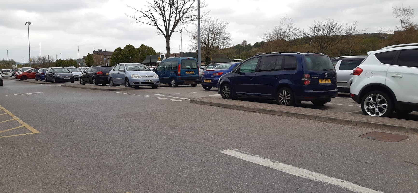 Busy car park with various parked vehicles, trees in the background, and cloudy skies overhead.
