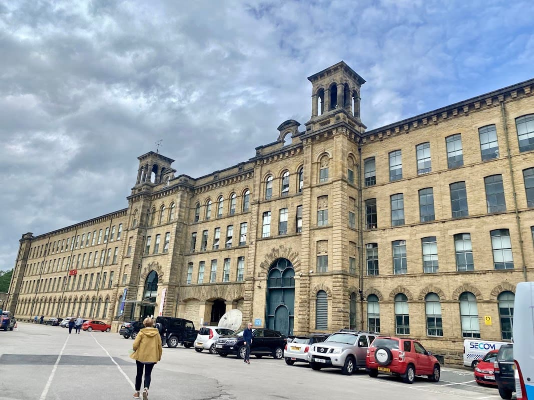 Historic stone building with large windows, a clock tower, and parked cars under a cloudy sky in Bradford.