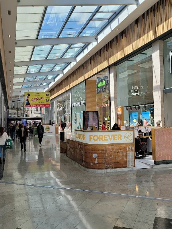 Bright indoor shopping area with shops, a café, and a skylight ceiling, bustling with people.