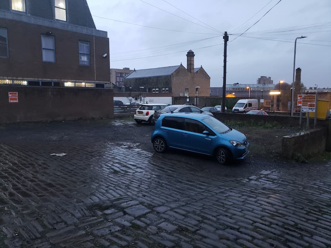 Cobblestone car park with parked cars, buildings in the background, and a cloudy sky in Bradford, Yorkshire.