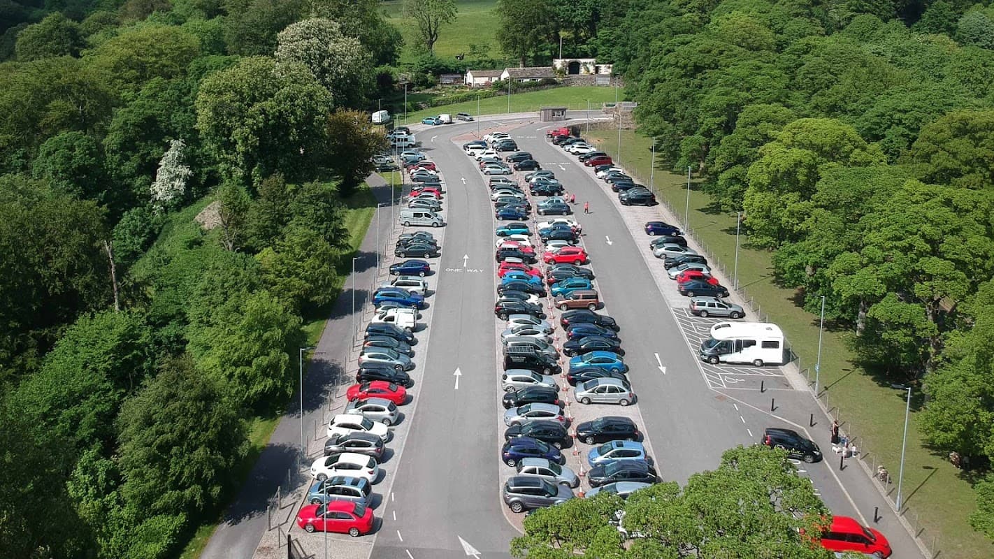 A busy pay and display car park filled with various parked cars, surrounded by lush greenery and trees.