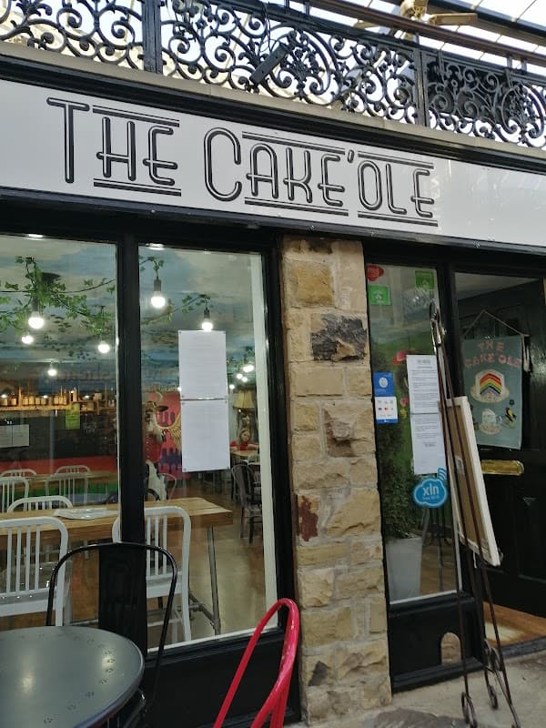 The Cake'ole café entrance with large windows, stone wall, and outdoor seating in a vibrant, welcoming atmosphere.