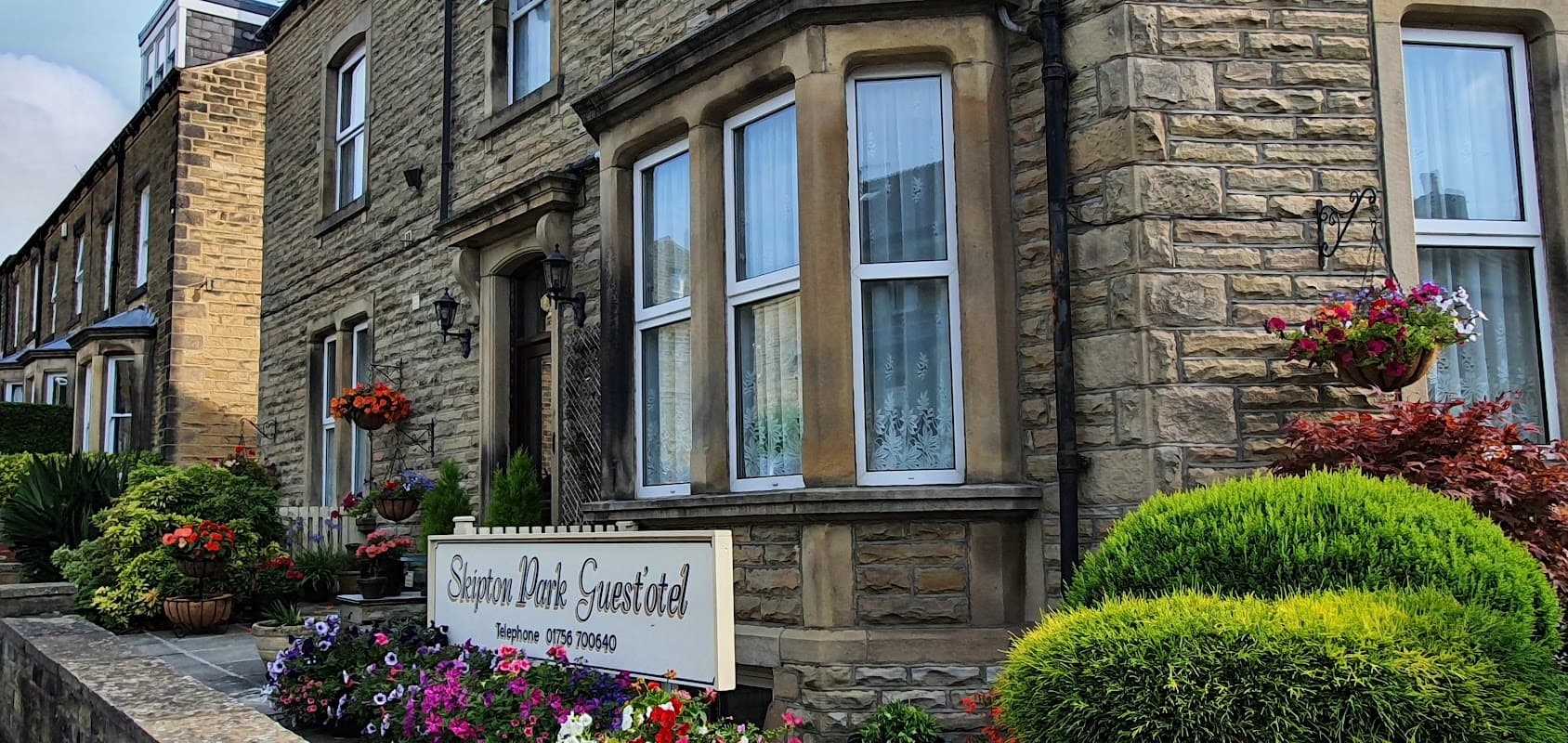 Stone building with large windows, colorful flower beds, and a sign for "Skipton Park Guest'otel" in Bradley, North Yorkshire.