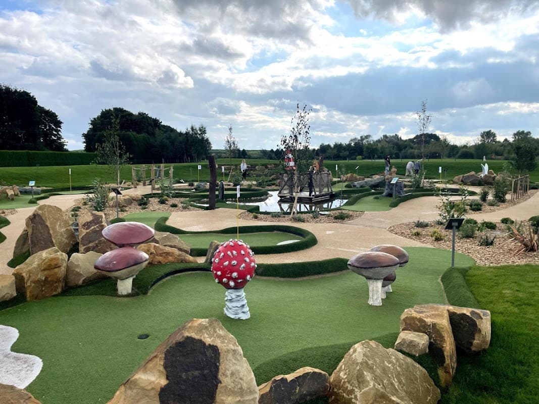 Colorful mini-golf course featuring whimsical mushrooms, rocks, and a pond, with players enjoying the game under a cloudy sky.