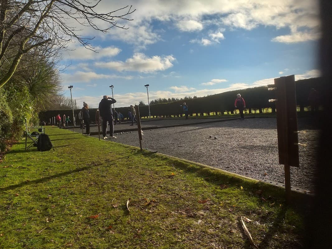 Petanque players on a gravel court under a bright sky, surrounded by greenery and wooden posts.