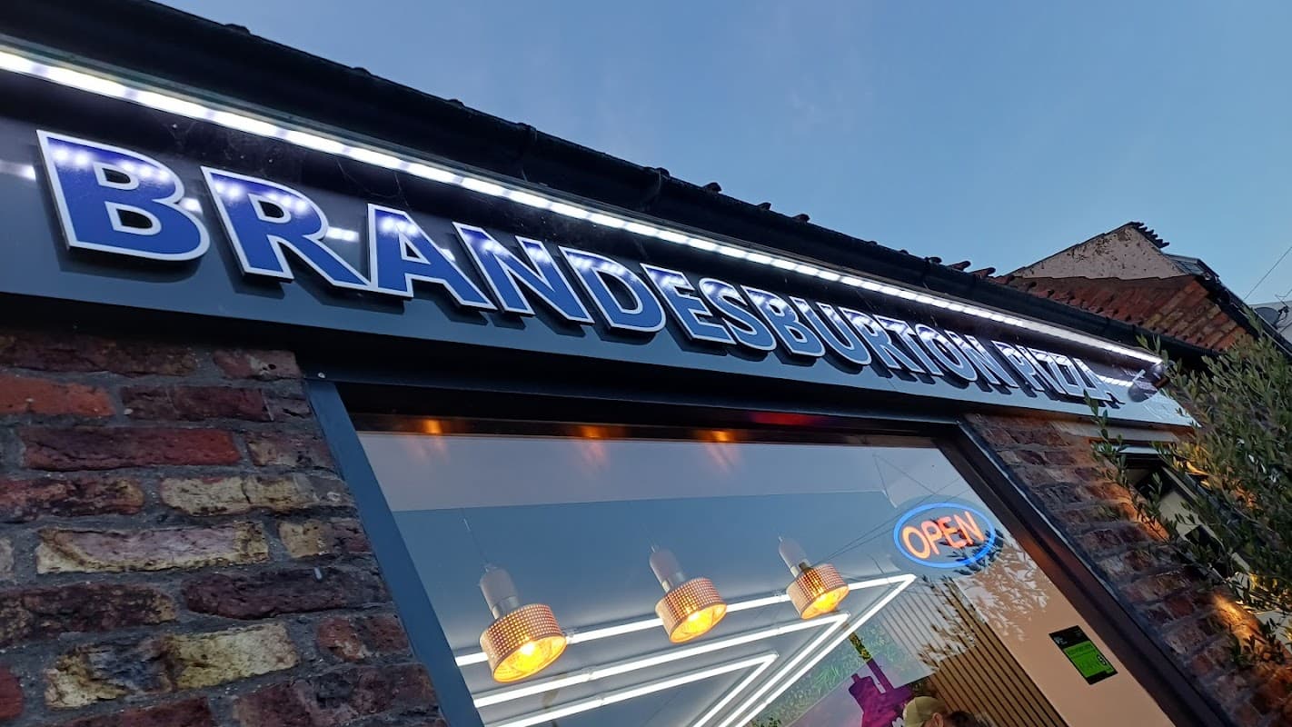 Sign for Brandesburton Pizza illuminated at dusk, featuring an "OPEN" sign and warm pendant lighting.