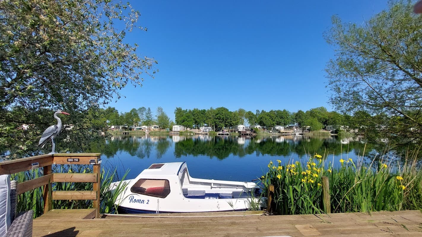 Serene lake view with a boat docked, surrounded by lush greenery and static caravans under a clear blue sky.