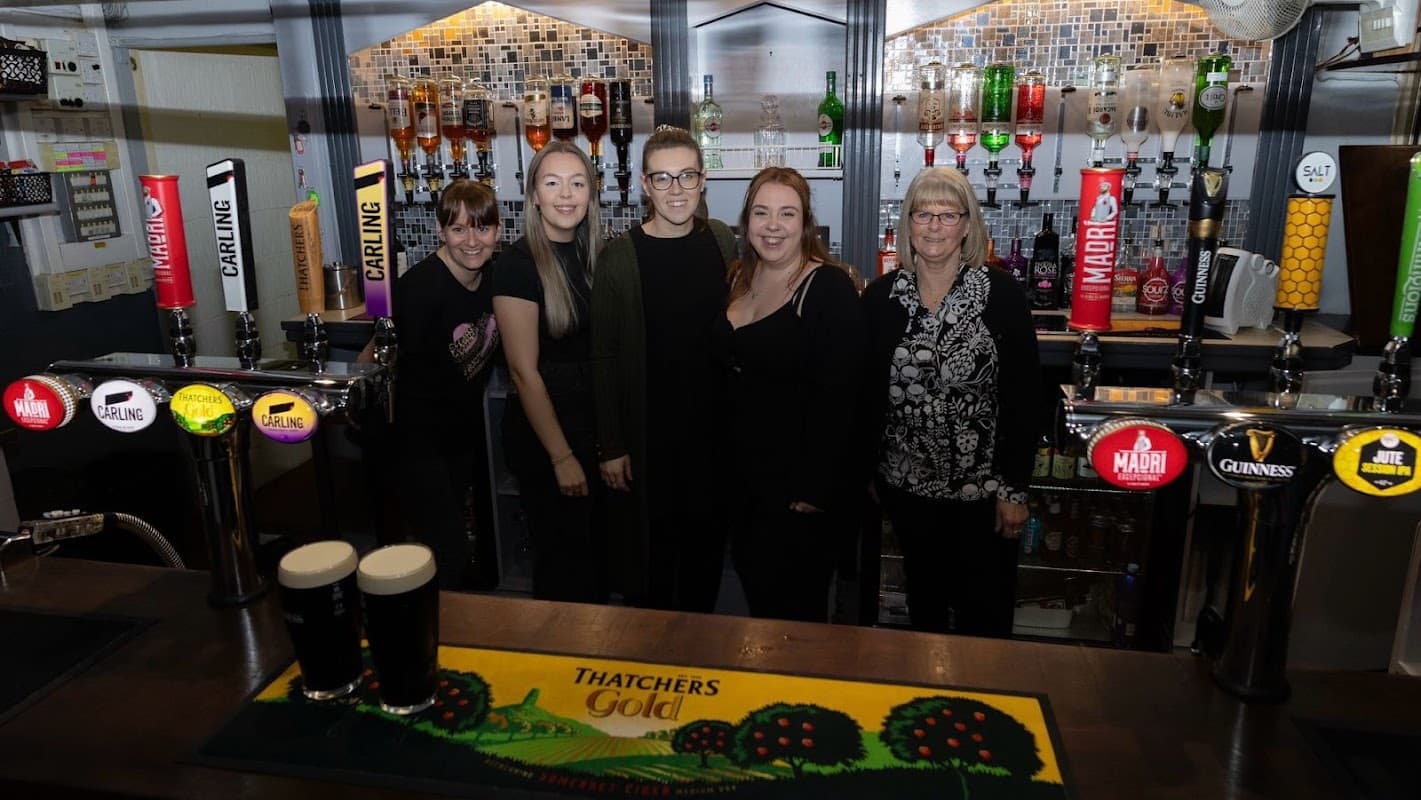 Five women stand behind a bar with various beer taps and a colorful logo mat featuring a countryside scene.