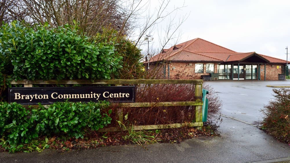 Brayton Community Centre sign with greenery and brick building in the background, cloudy sky above.