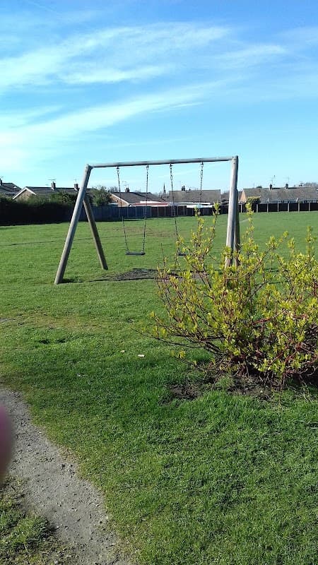 Swing set on a grassy field with a bush in the foreground and houses visible in the background under a clear blue sky.