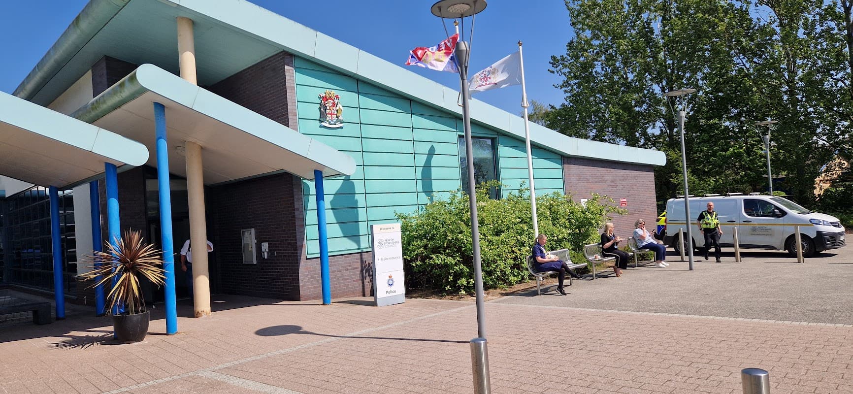 Modern building with teal panels, flags, and people sitting on benches outside the North Yorkshire Council Selby Offices.