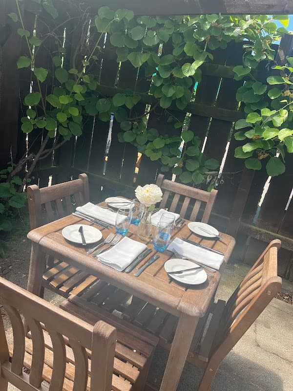Wooden table set for dining with plates, cutlery, and a flower vase, surrounded by greenery in a cozy outdoor setting.