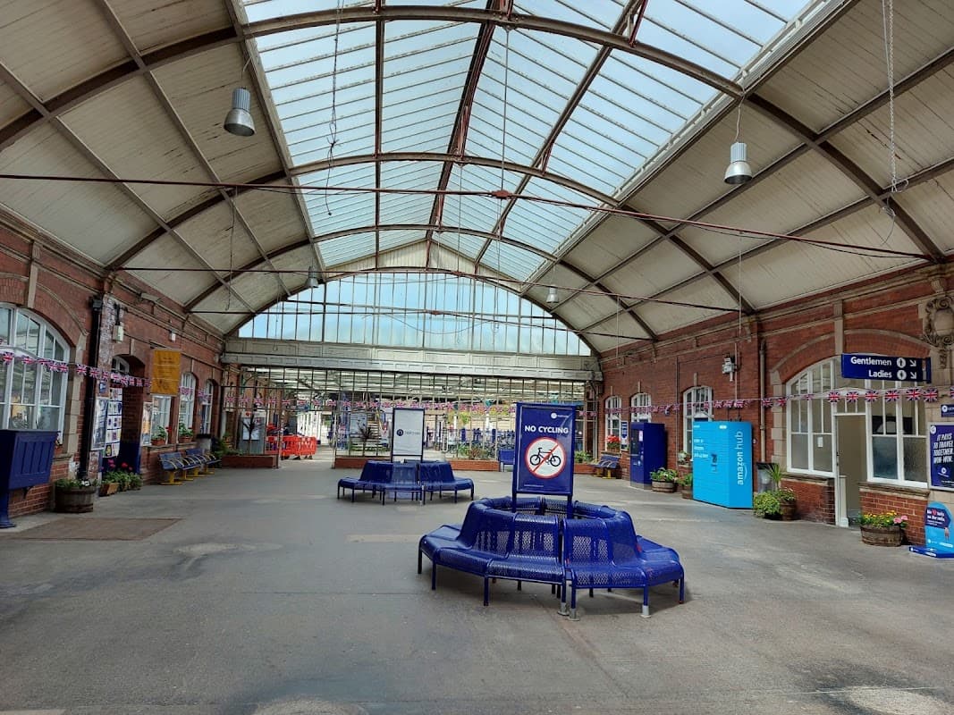 Spacious railway station interior with high glass roof, blue seating, and signage, featuring colorful flower planters.
