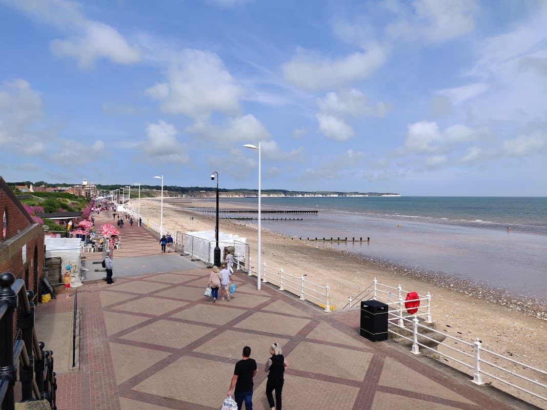 Car park near the beach, with a promenade, sandy shore, and distant cliffs under a partly cloudy sky.