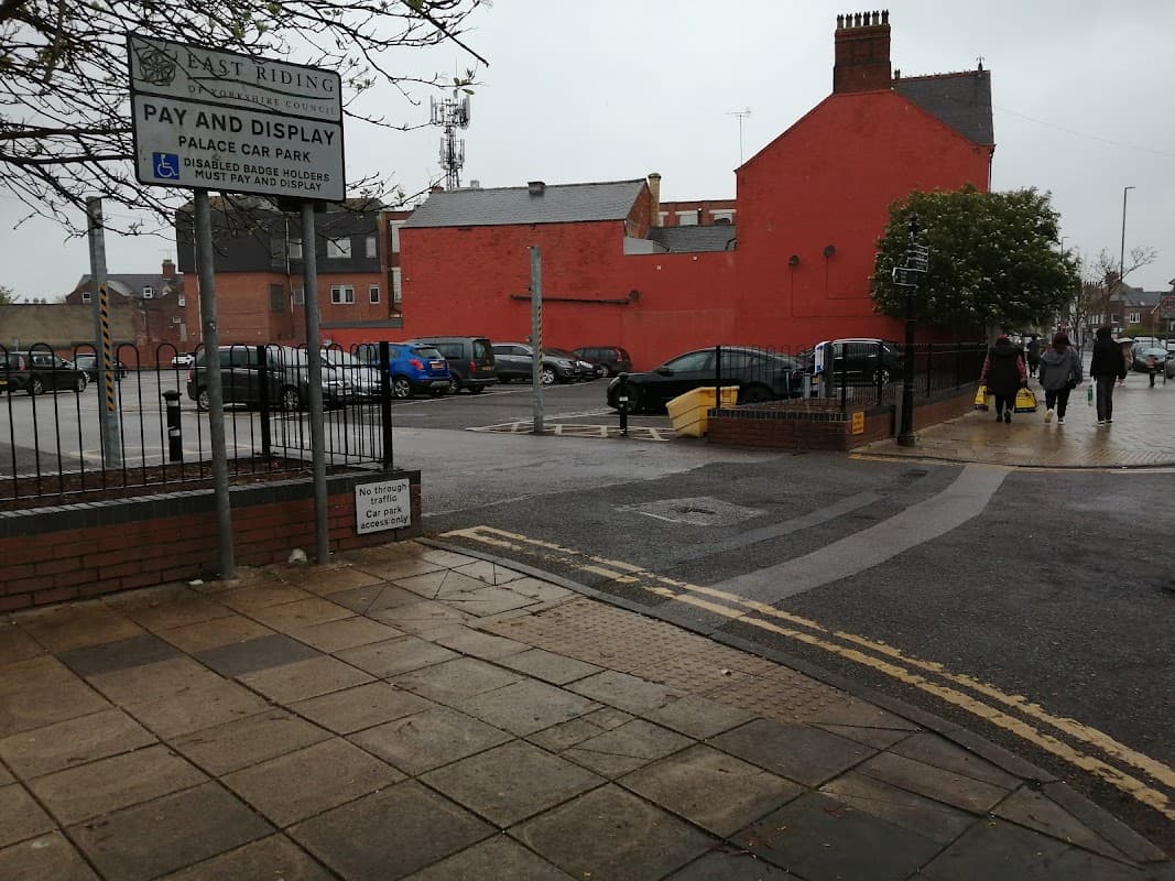 Pay & Display sign at Palace Car Park, with parked cars, a red building, and pedestrians walking in rainy weather.