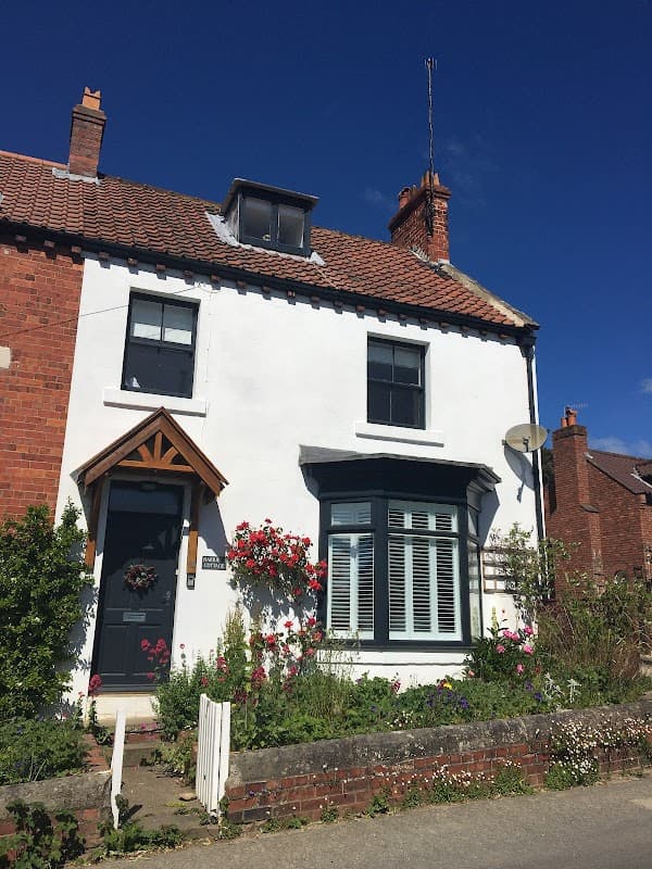 White and red brick cottage with a wooden door, flower-filled garden, and blue sky in Briggswath, Yorkshire.