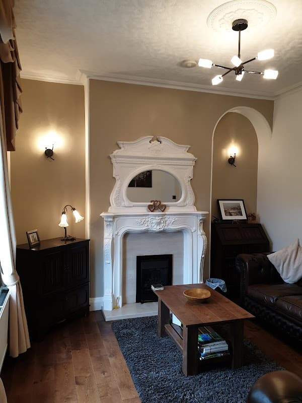 Cozy living room with a white fireplace, mirror, wooden table, leather sofa, and warm lighting.