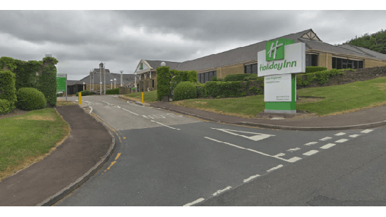 Entrance to Holiday Inn with green signage, surrounded by neatly trimmed hedges and a cloudy sky in Brighouse, Yorkshire.