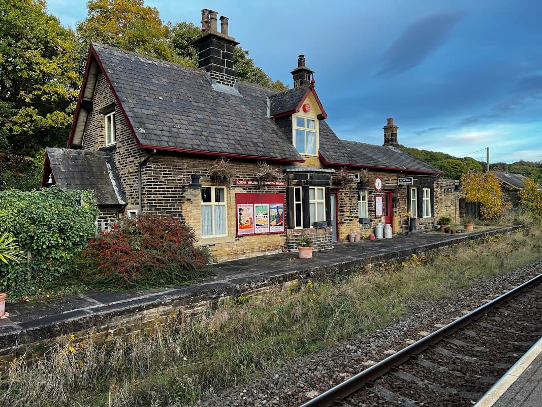 Historic railway station building with stone walls, slate roof, and surrounding greenery, set against a dramatic sky.