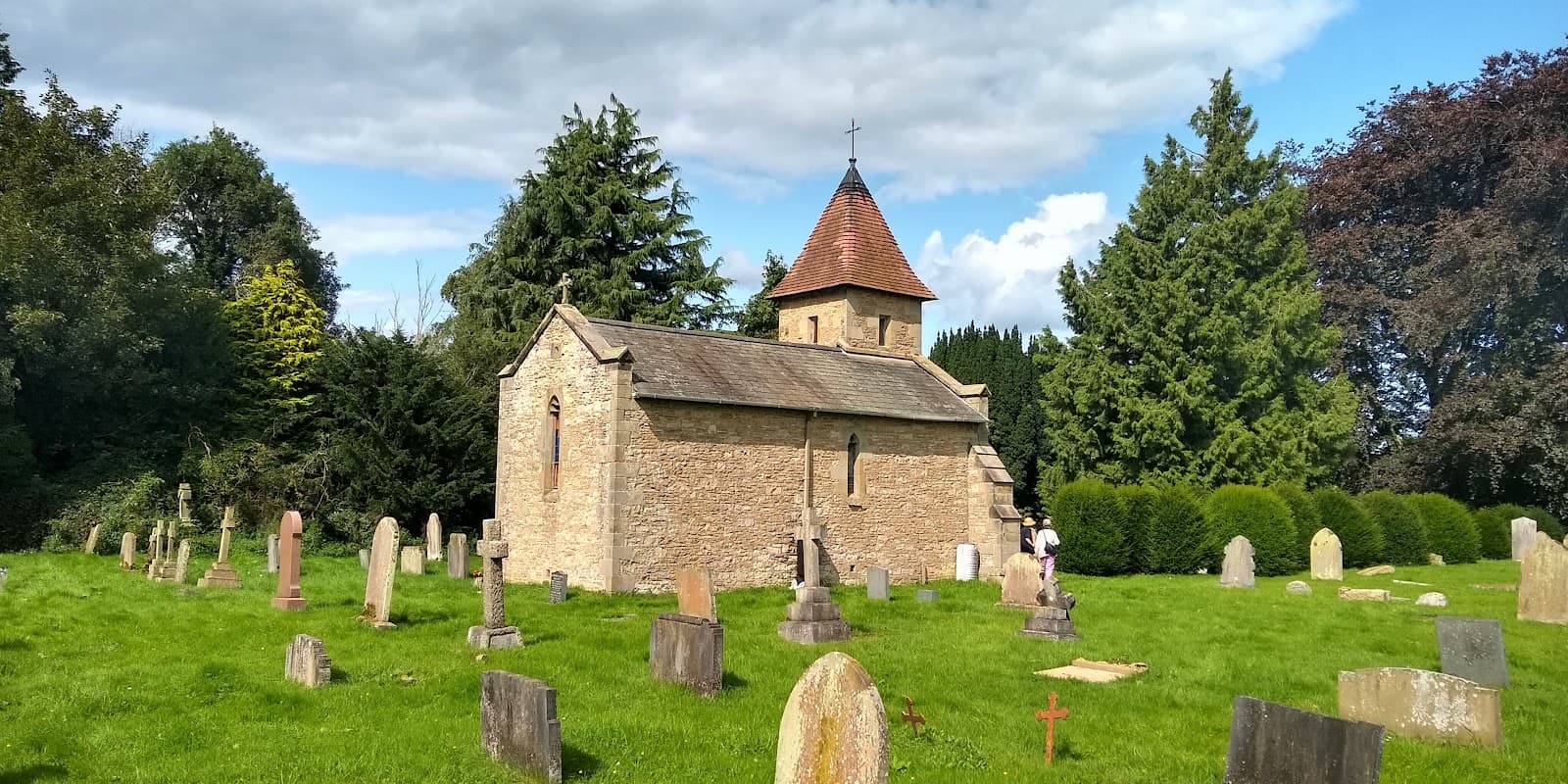 Chapel of Rest surrounded by gravestones and lush greenery under a partly cloudy sky in Brompton-by-Sawdon.