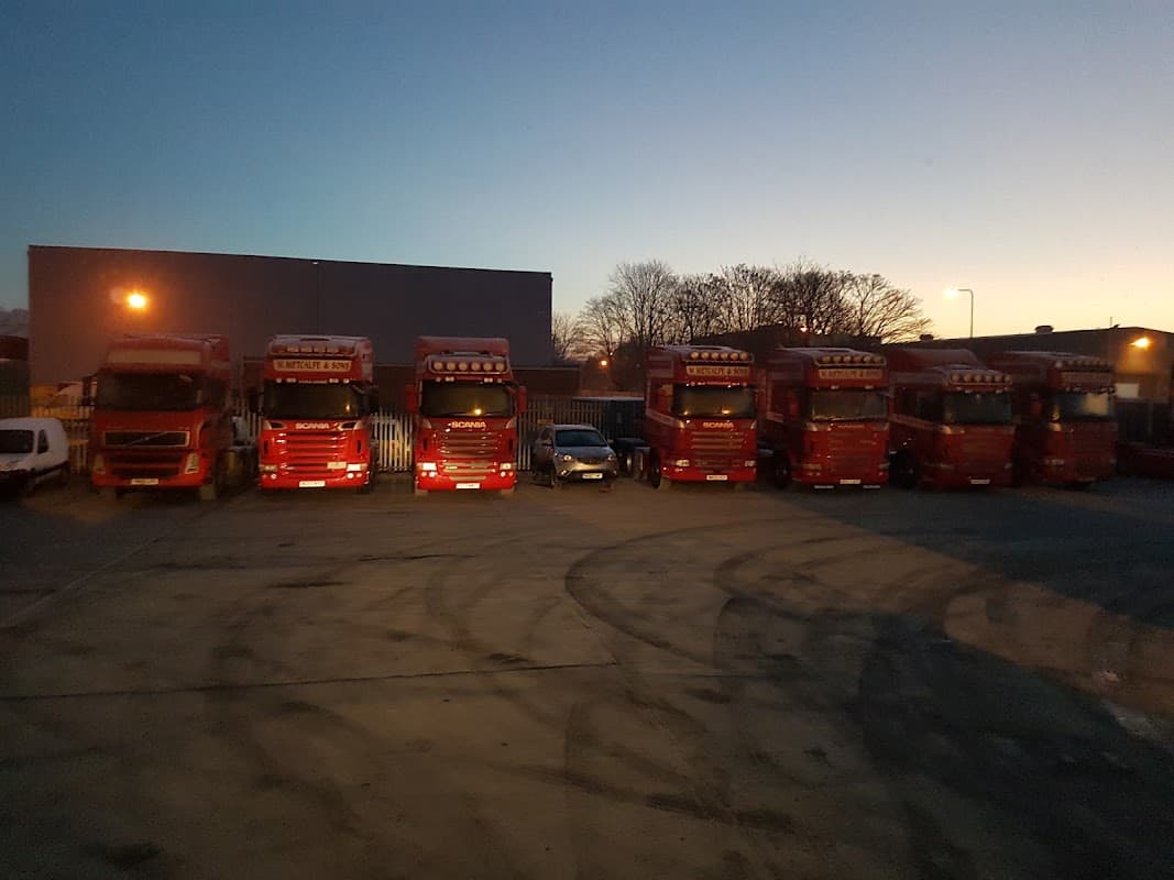 W Metcalfe & Sons Ltd trucks lined up in a yard at dusk, with a few parked vehicles and trees in the background.