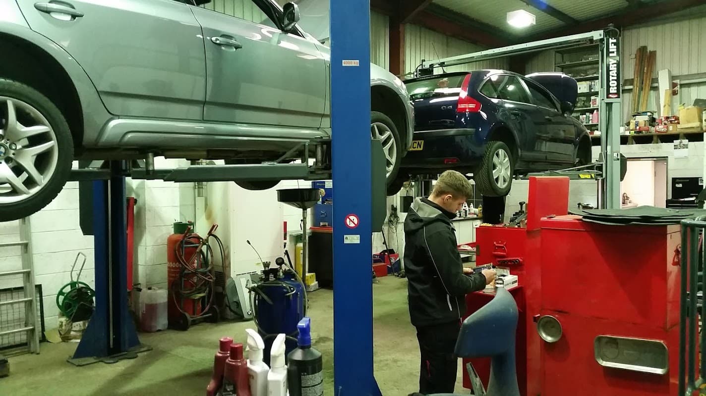 Two cars on lifts in a garage, with a mechanic working at a red workstation surrounded by tools and equipment.