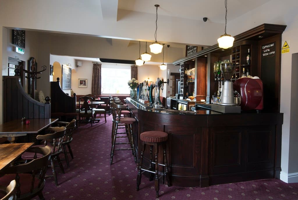 Cozy bar interior with wooden counter, stools, and a warm atmosphere, featuring drinks on display and soft lighting.