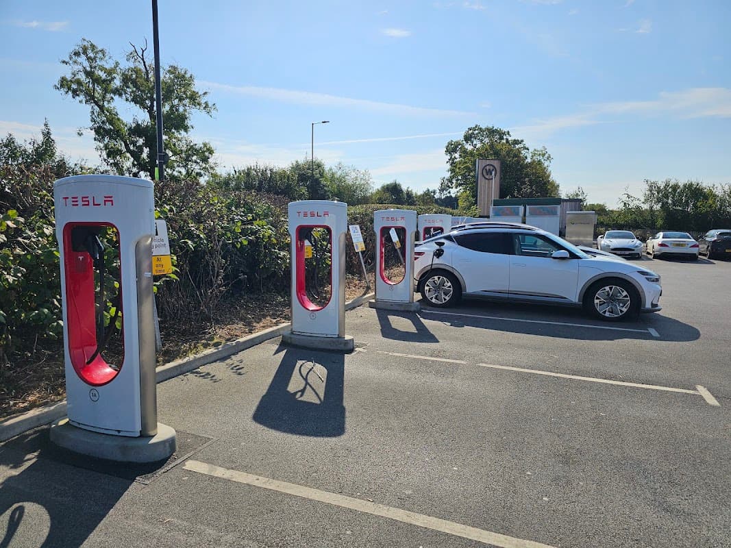 Tesla Supercharger station with charging units and a parked Tesla vehicle in a sunny parking lot.