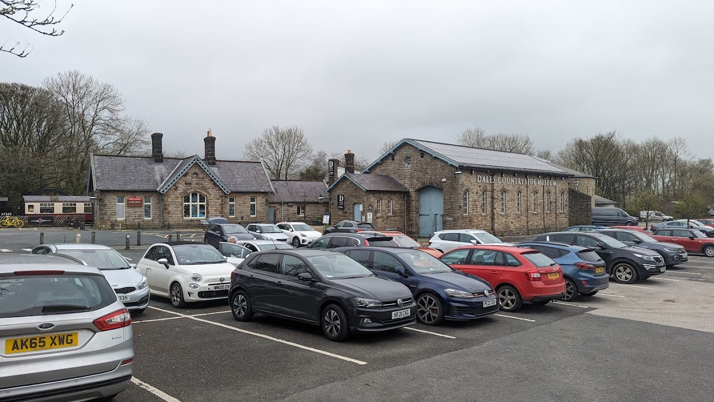 Stone building with a slate roof, surrounded by parked cars and trees, under a cloudy sky.