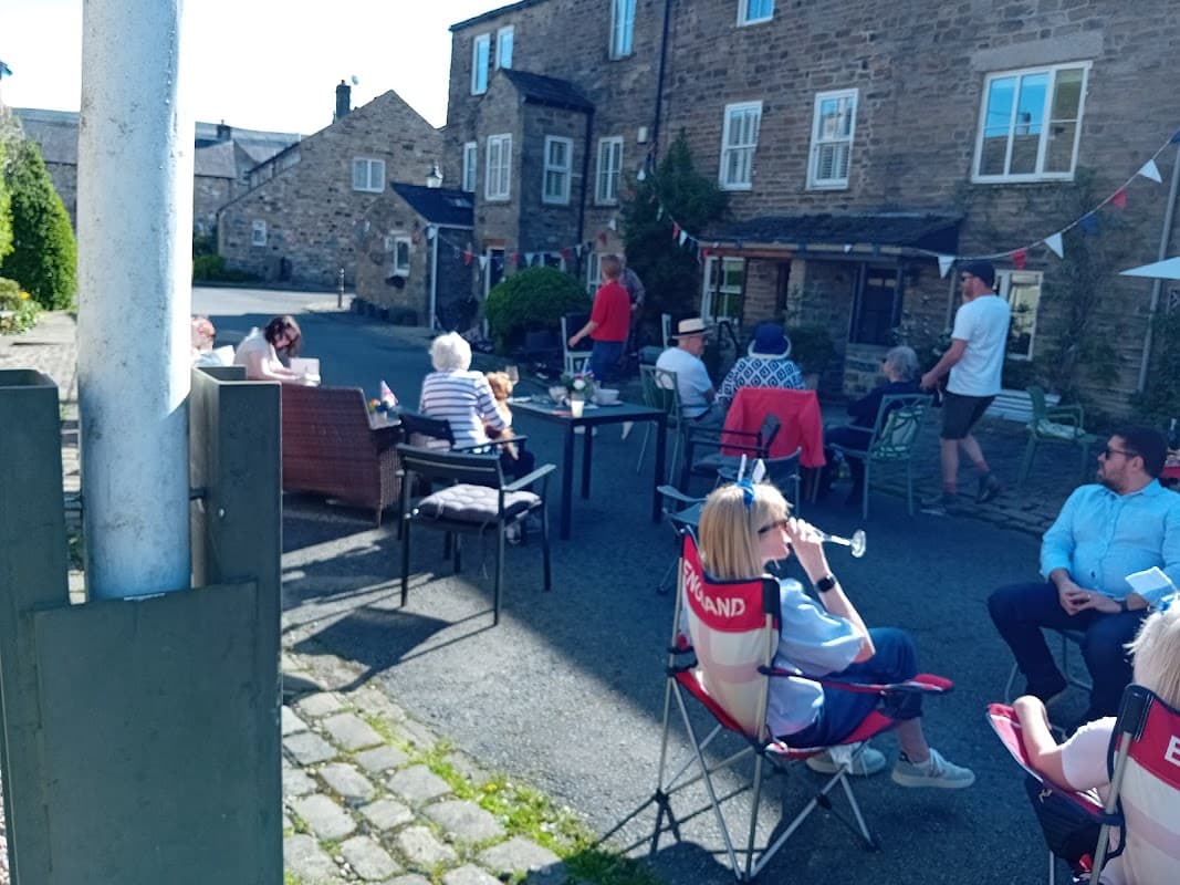 People socializing outside at Burton Leonard Cricket Club, with chairs, tables, and festive bunting decorating the area.