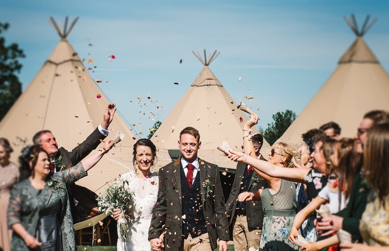 A joyful couple walks through confetti, surrounded by smiling guests and teepee tents at Aldby Park Weddings.