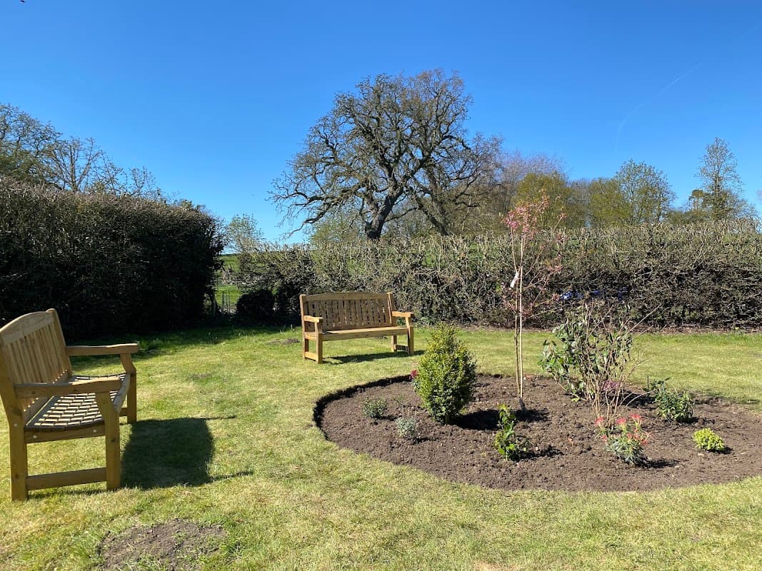 A sunny community garden with wooden benches, a circular flower bed, and lush greenery in Buttercrambe, Yorkshire.