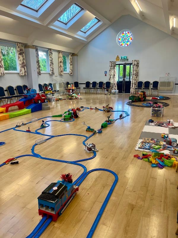 Bright, spacious village hall with train tracks, toys, and colorful seating, featuring large windows and a stained glass window.