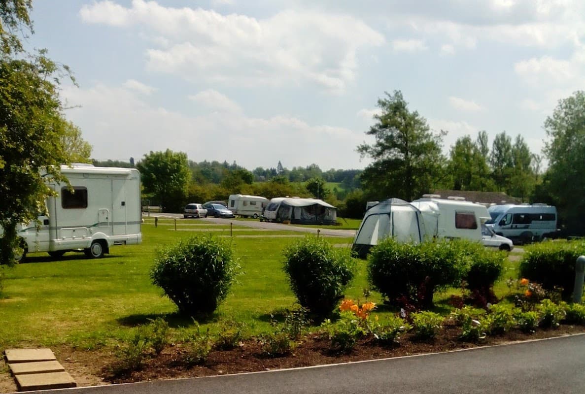 Caravans parked on a grassy area, surrounded by trees and flower beds under a partly cloudy sky.