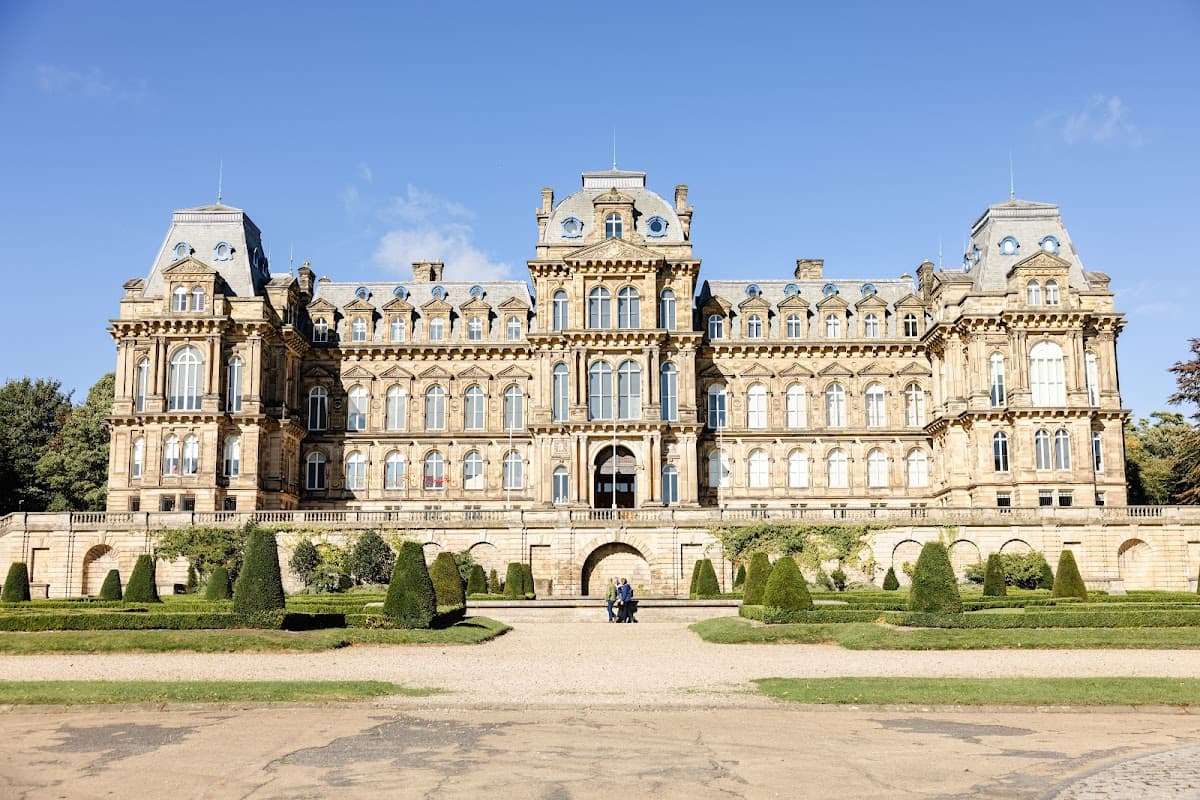 Victorian mansion with ornate architecture, surrounded by manicured gardens and trees under a clear blue sky.