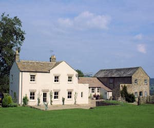 White farmhouse with stone chimney, adjacent rustic stone barn, set in lush green landscape under blue sky.