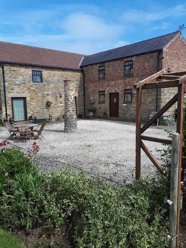 Holiday cottages with stone walls, gravel courtyard, wooden seating, and lush greenery under a blue sky in Yorkshire.