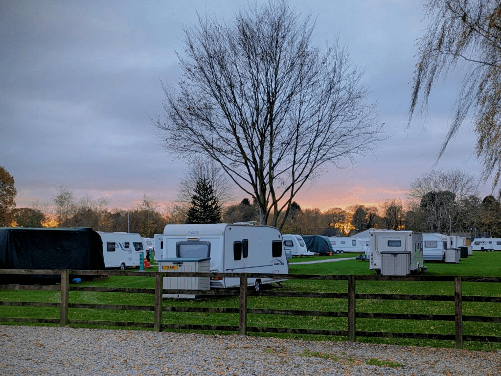 Caravans lined up on green grass, with trees and a sunset in the background at Carlton Miniott Holiday Park.
