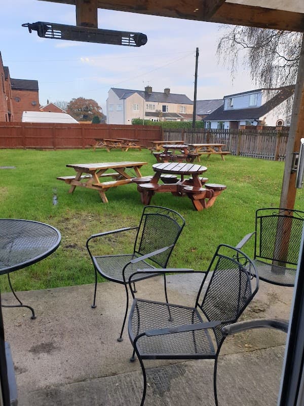 Outdoor seating area with picnic tables on green grass, surrounded by wooden fencing and residential buildings in the background.