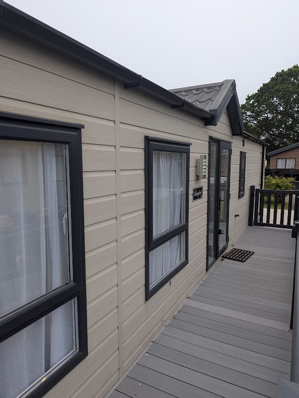 Exterior view of a holiday park cabin with gray siding, windows, and a wooden deck in a green, tree-lined setting.