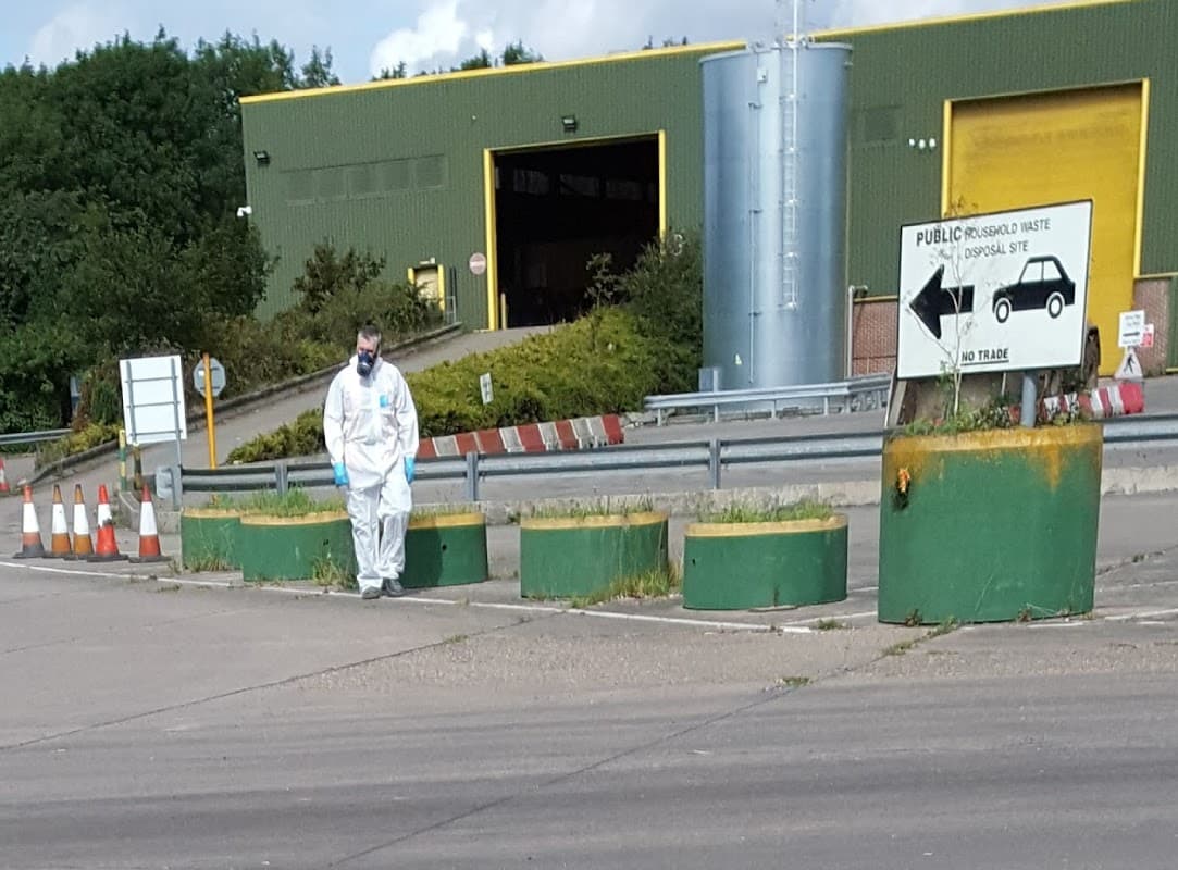 Household waste and recycling site with green barriers, signage, and a worker in protective gear walking on the pavement.