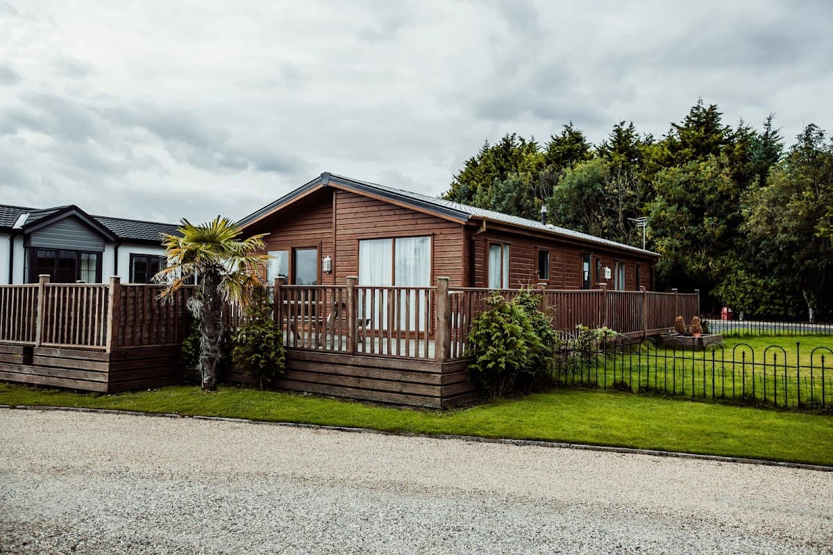 Wooden lodge with a deck, surrounded by greenery and a gravel path, set against a cloudy sky in Little Eden Country Park.