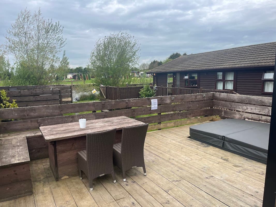 Wooden deck with a table and chairs, overlooking a tranquil pond and lodge, surrounded by greenery under a cloudy sky.