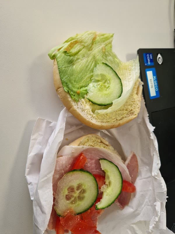 A sandwich bun topped with lettuce, cucumber, and sliced ham with tomatoes, resting on crumpled white paper.