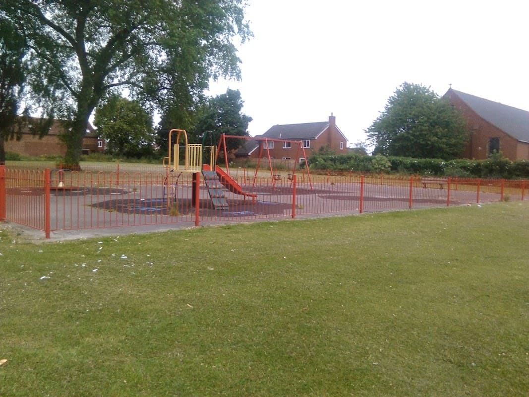 Playground with swings and slides, surrounded by a red fence and green grass, near residential houses in Castleford.