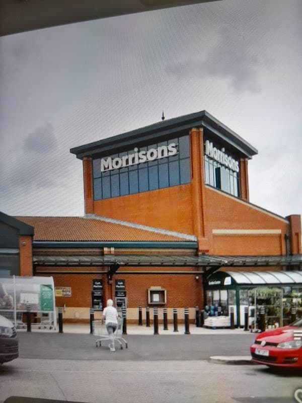 Morrisons supermarket with a shopper pushing a cart, surrounded by parking spaces and a cloudy sky.