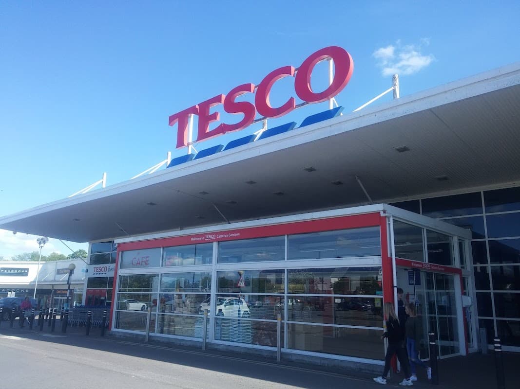 Tesco Superstore exterior with large red signage, glass entrance, and shoppers outside in Catterick Garrison, Yorkshire.